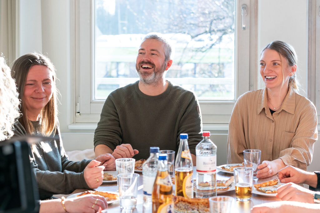 Mehrere Mitarbeitende von RBM Ritzmann + Dr. Blödorn sitzen zusammen am Tisch, lachen und unterhalten sich; auf dem Tisch stehen Getränke und Essen – kollegiale Atmosphäre in der Steuerberatung und Wirtschaftsprüfung.