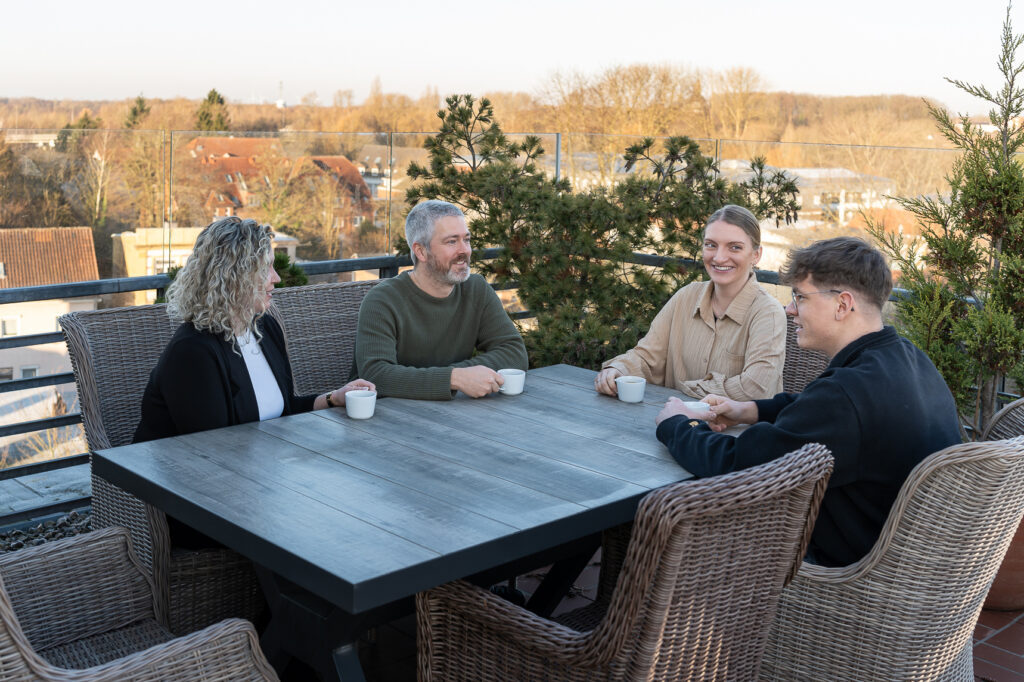 Vier Mitarbeitende von RBM Ritzmann + Dr. Blödorn sitzen an einem Tisch auf der Dachterrasse und sprechen miteinander; auf dem Tisch stehen Tassen – Teamgespräch in der Steuerberatung und Wirtschaftsprüfung.