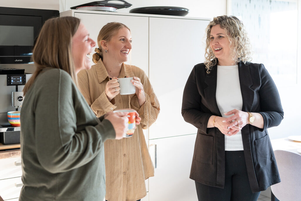 Drei Mitarbeiterinnen von RBM Ritzmann + Dr. Blödorn stehen in der Büroküche, lachen und sprechen miteinander; zwei halten Tassen – Einblick in die Teamkultur der Steuerberatung und Wirtschaftsprüfung.